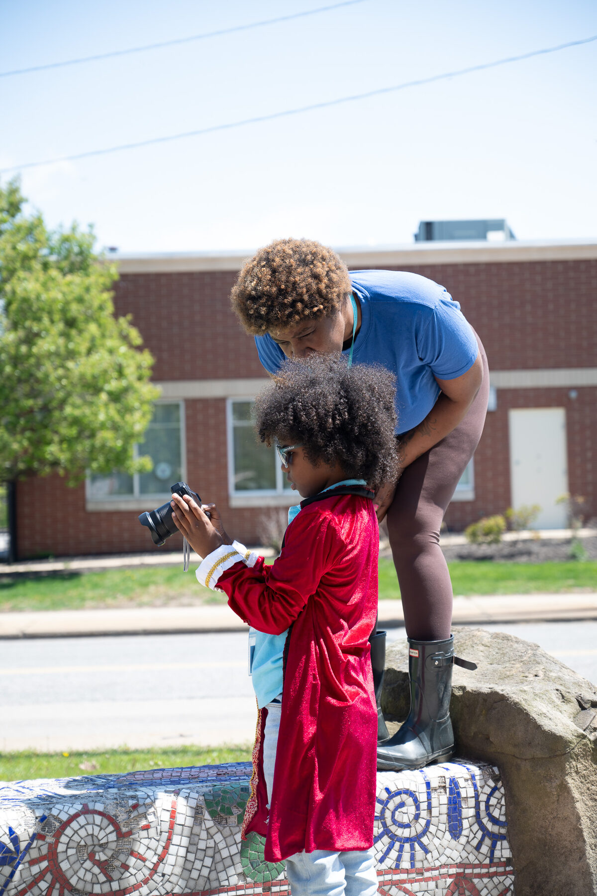 An adult helps a young artist in a red velvet coat review photos on a camera outdoors beside a mosaic bench